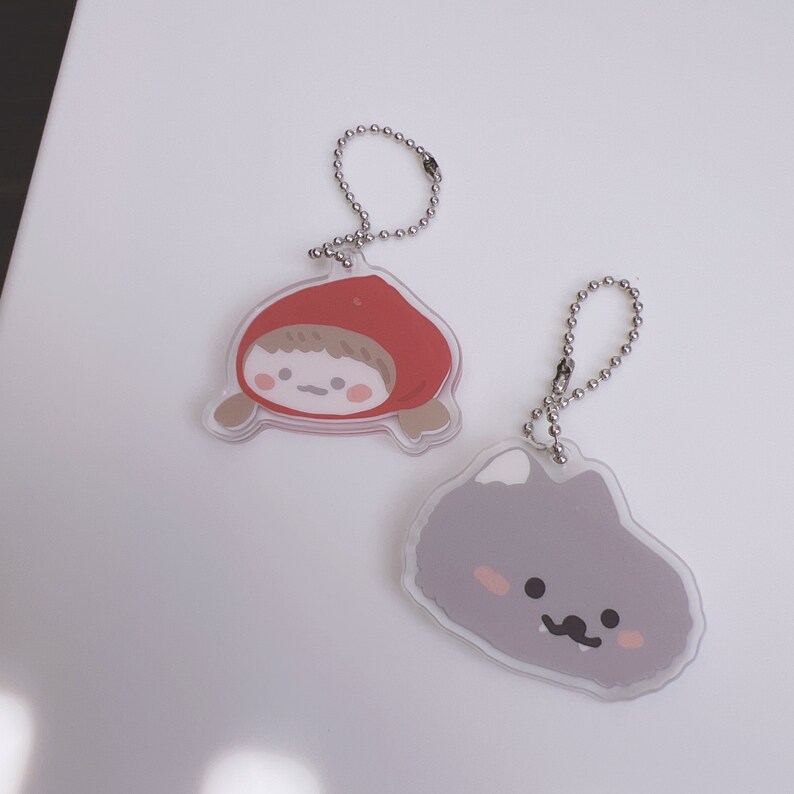 Op de afbeelding: Twee transparante acryl sleutelhangers met cartoonfiguren. De ene sleutelhanger heeft een meisje met rood haar dat een rode kapmantel draagt. De andere sleutelhanger heeft een grijs, pluche wezen met een wit oor en een snor.