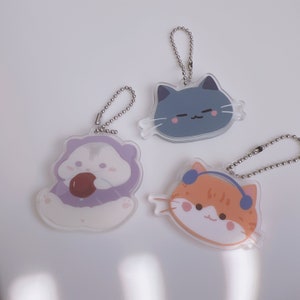 Op de afbeelding: Drie schattige dieren sleutelhangers met zilveren kettingen. De eerste sleutelhanger heeft een hamster in een paarse outfit die een bruine traktatie vasthoudt. De tweede sleutelhanger heeft een blauwe kat met roze wangen. De derde sleutelhanger heeft een oranje kat met blauwe koptelefoon.