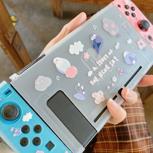 K&ouml;nnte beinhalten: Eine graue Nintendo Switch-Konsole mit einer Schutzh&uuml;lle mit einem skurrilen Design. Die H&uuml;lle ist mit Illustrationen von V&ouml;geln, Wolken und Blumen sowie dem Text "A Story of the Blue Sky" verziert. Die Controller sind rosa und blau.