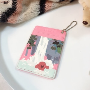 May include: A rectangular pink card holder featuring a graphic illustration of a waterfall, green trees, and a small red character in the water. The holder includes a silver metal ball chain attached to the top right corner for hanging.