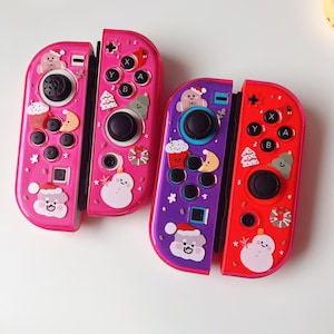 May include: Two colorful game controllers with a festive holiday theme. One is pink, the other is purple and red. Each controller has a black joystick and buttons with holiday-themed stickers, including snowmen and Santa hats.