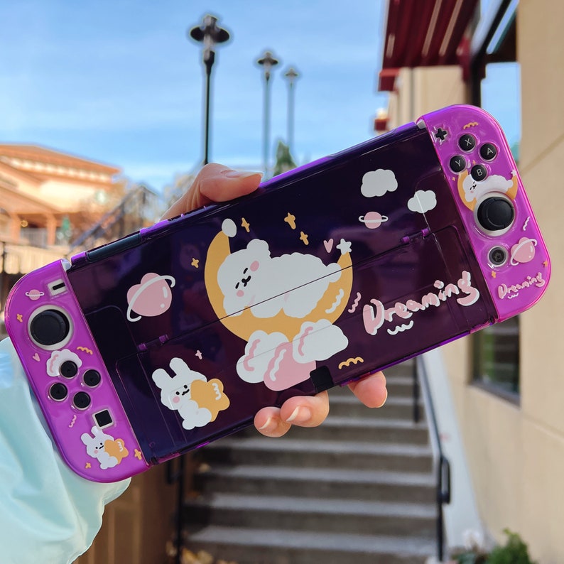 May include: Purple Nintendo Switch case with a clear back panel featuring a cartoon bear sleeping on a crescent moon with clouds and stars. The text "Dreaming" is printed on the case.