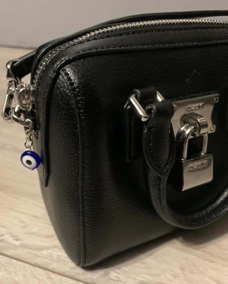 May include: A black leather handbag with a silver lock and chain strap. A blue and white evil eye charm hangs from the strap.