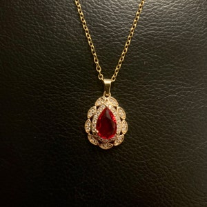 May include: A gold-toned necklace featuring a teardrop-shaped pendant with a central red gemstone surrounded by a halo of small clear stones. The pendant is suspended from a delicate gold-toned chain with oval-shaped links.