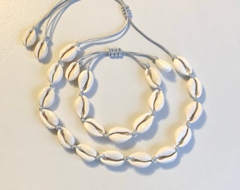Op de afbeelding: Een set van twee schelpenkettingen met grijze koorden. De kettingen zijn gemaakt van witte kauri-schelpen en hebben verstelbare koorden.