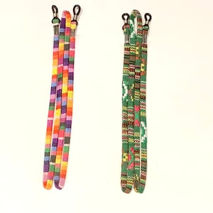May include: Two pairs of spectacle straps. One pair has a colourful pattern of orange, pink, yellow, purple, and blue squares. The other pair has a green, brown, and red geometric design. Both have black rubber loops and silver metal connectors.