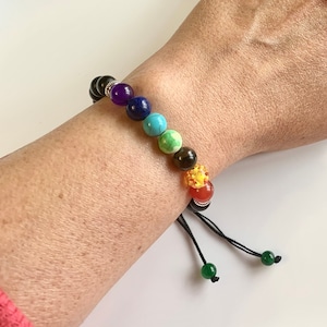 May include: A black string bracelet with seven colourful beads representing the seven chakras. The beads are purple, blue, turquoise, green, brown, orange, and red. The bracelet has a green bead on each end of the string.