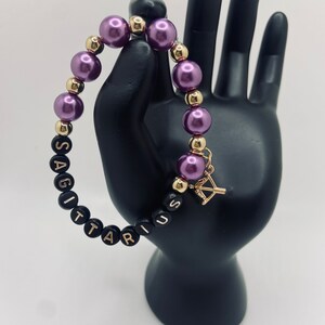 May include: Purple and gold beaded bracelet with the word "Sagittarius" spelled out in black beads. The bracelet has a gold arrow charm.