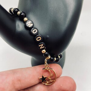 May include: Black beaded bracelet with gold accents and the word "MOON" spelled out in letter beads. A gold charm with a crescent moon and star dangles from the bracelet. The bracelet is held against a black background.