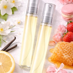 May include: Two clear glass perfume bottles filled with yellow liquid, with silver sprayers. The bottles are surrounded by white flowers, a lemon slice, vanilla beans, strawberries, a honeycomb, and pink macarons.