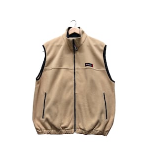 Puede incluir: Un chaleco de forro polar de color beige con una cremallera negra y ribetes negros. El chaleco tiene un pequeño logotipo rojo y negro en la parte delantera que dice "All-Terrain".