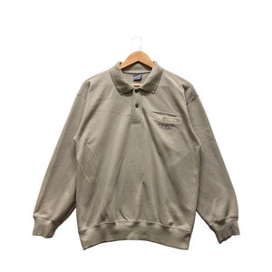 Peut inclure: Un polo à manches longues de couleur beige clair avec une petite poche sur la poitrine gauche. La poche comporte un petit logo brodé qui dit "ODYSSEY".