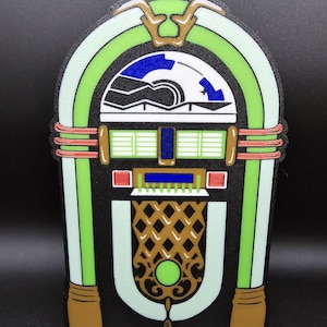 May include: A retro-style jukebox design, measuring 9.5 x 5.75 inches. The image features a black background and a colorful jukebox with green, white, brown, red, and blue accents. The jukebox has a detailed speaker grill and a classic design.