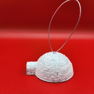 May include: A white igloo ornament with a silver hanging loop. The igloo has a textured, snowy appearance and is set against a red background. This festive decoration is perfect for winter holiday themes.