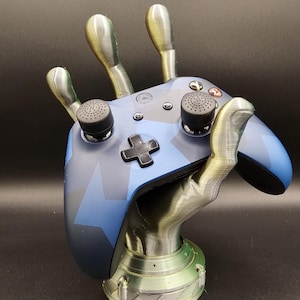 May include: A 3D printed hand holding a blue and black video game controller. The hand is made of a light gray plastic and has four fingers. The controller is a standard Xbox controller with a blue and black camouflage pattern. The hand is holding the controller in a relaxed position, as if it is about to play a game.