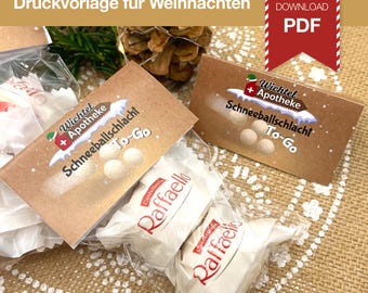 Op de afbeelding: Close-up van verpakte traktaties met de tekst "Wichtel + Apotheke Schneeballschlacht To-Go" op een bruine kaart. De afbeelding bevat ook Raffaello-snoepjes en de tekst "DIGITALER DOWNLOAD PDF".