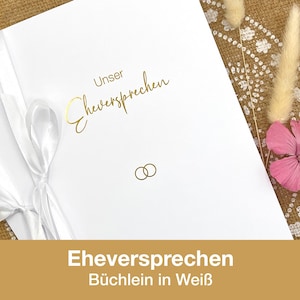 Könnte beinhalten: Ein weißes Hochzeitsgelübdebuch mit goldfarbenem Schriftzug "Unser Eheversprechen" und zwei goldenen Eheringen. Auf der linken Seite ist ein weißes Band gebunden. Der Text "Eheversprechen Büchlein in Weiß" befindet sich unten.