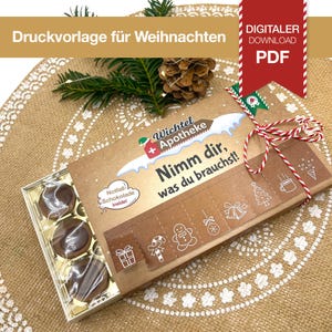 Pode incluir: Uma caixa de cartão castanha com o texto "Nimm dir, was du brauchst!" e "Wichtel Apotheke" impressos. A caixa contém bombons de chocolate e é decorada com ilustrações de Natal. Uma corda vermelha e branca está amarrada em volta da caixa.