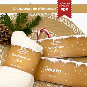 Pode incluir: Um par de meias brancas de malha com uma faixa de papel castanho com o texto "Kuschelsocken Zeit" e flocos de neve. A imagem também inclui uma bengala de doces e um pinha, sugerindo um tema de férias.