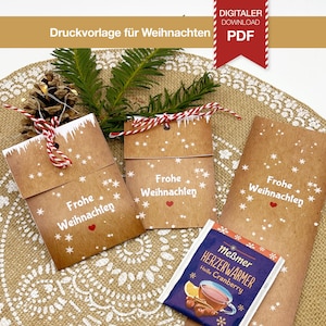 Op de afbeelding: Kraftpapieren cadeaulabels en verpakkingen met de woorden "Frohe Weihnachten" en sneeuwvlokontwerpen. Een theezakje met de woorden "Meßmer Herzerwärmer Heile Cranberry" is ook zichtbaar. De afbeelding bevat ook een dennenappel en groen.