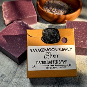 May include: Handcrafted soap bars and a small package with a black wax seal. The package has a label that reads "SAVAGEMOON SUPPLY Elixir HANDCRAFTED SOAP". The soap bars are a deep purple color, and a wooden bowl with dried lavender is in the background.