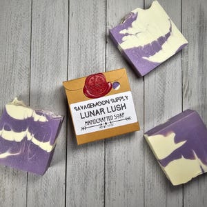 May include: Three bars of handmade soap with a purple and white swirl design. The soap is packaged in a brown box with the text "Savagemoon Supply Lunar Lush Handcrafted Soap Net Wt. 4.5 oz."