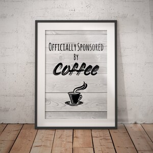 Könnte beinhalten: Ein gerahmter Druck mit weißem Holzmaserungshintergrund und schwarzem Text, der "Offiziell gesponsert von Kaffee" lautet, mit einer stilisierten Kaffeetassenillustration.