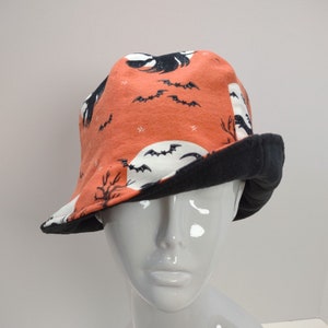 Puede incluir: Un gorro de forro polar negro y naranja con un estampado de Halloween con murciélagos y lunas. El gorro tiene un forro de polar negro.