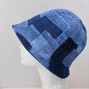 May include: A blue denim bucket hat with a patchwork design. The hat features various shades of blue denim fabric, creating a textured and visually interesting pattern. The hat is displayed on a white mannequin head.