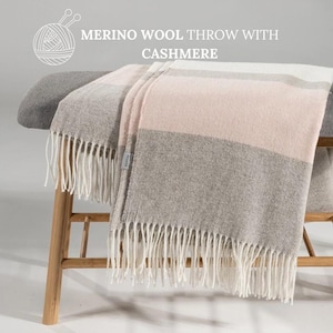 Op de afbeelding: Een zachte, grijs en roze gestreepte deken met witte franjes. De deken is gemaakt van merinowol en kasjmier en is over een houten bank gelegd. De tekst "MERINO WOOL THROW WITH CASHMERE" staat boven de deken.