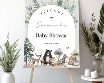 May include: Welcome to Savannah's Baby Shower sign with a woodland theme. The sign features a green background with a circle that says "Welcome to Savannah's Baby Shower" and a date. The circle is surrounded by green leaves and branches. Below the circle, there are cartoon animals in a forest setting, including a bear, deer, fox, raccoon, owl, and squirrel.