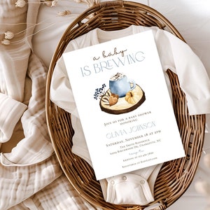 Op de afbeelding: Een witte uitnodigingskaart voor een babyshower met de tekst 'a baby IS BREWING' boven een aquarelillustratie van een blauwe mok, een pompoen en bessen op een houten schijf. De kaart bevat de evenementgegevens voor Olivia Johnson.