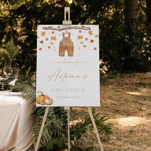 May include: A wooden easel displays a baby shower sign with the text "Welcome to Autumn's Baby Shower November 15, 2024." The sign features watercolor illustrations of overalls, pumpkins, and autumn leaves. The scene is set outdoors.