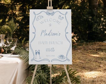 May include: A white welcome sign with blue text and illustrations. The sign reads "Welcome to Madison's Baby Brunch, April 12, 2020". The sign is on a wooden easel in a forest setting.