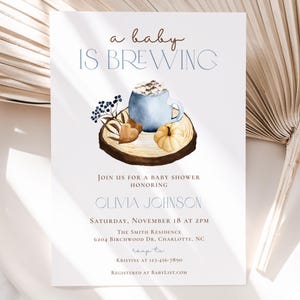 Op de afbeelding: Een witte uitnodigingskaart voor een babyshower met de tekst 'a baby IS BREWING' boven een aquarelillustratie van een blauwe mok met marshmallows, een kleine pompoen en bessen op een houten schijf. De kaart bevat details voor het evenement.
