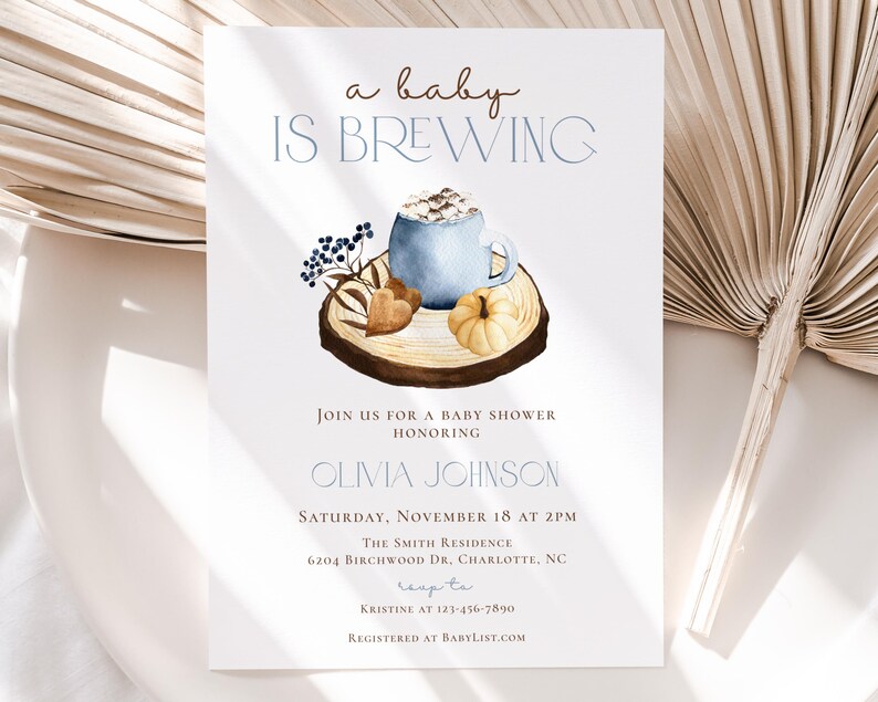 Op de afbeelding: Een witte uitnodigingskaart voor een babyshower met de tekst 'a baby IS BREWING' boven een aquarelillustratie van een blauwe mok met marshmallows, een kleine pompoen en bessen op een houten schijf. De kaart bevat details voor het evenement.