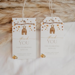 May include: Two white baby shower thank you tags. Each tag features a watercolor illustration of brown overalls hanging from a branch with autumn leaves. The text reads "thank YOU FOR CELEBRATING WITH US!" and includes the baby shower date.