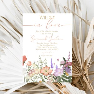May include: A white bridal shower invitation with gold lettering that reads "WILDLY in love" and features a watercolor illustration of a bouquet of pink, purple, and orange flowers. The invitation includes the bride's name, the date and time of the shower, the location, and RSVP information.