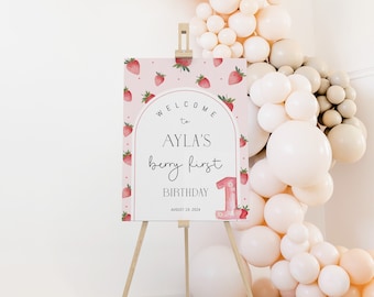 May include: A pink welcome sign with a strawberry pattern and the text "Welcome to Ayla's Berry First Birthday August 18, 2024". The sign is on a wooden easel.