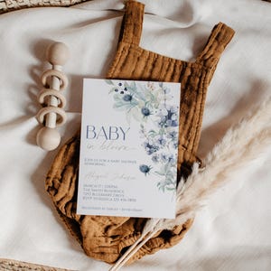 a baby shower invitation with a floral design, placed on a white cloth with baby toys and pom-poms.