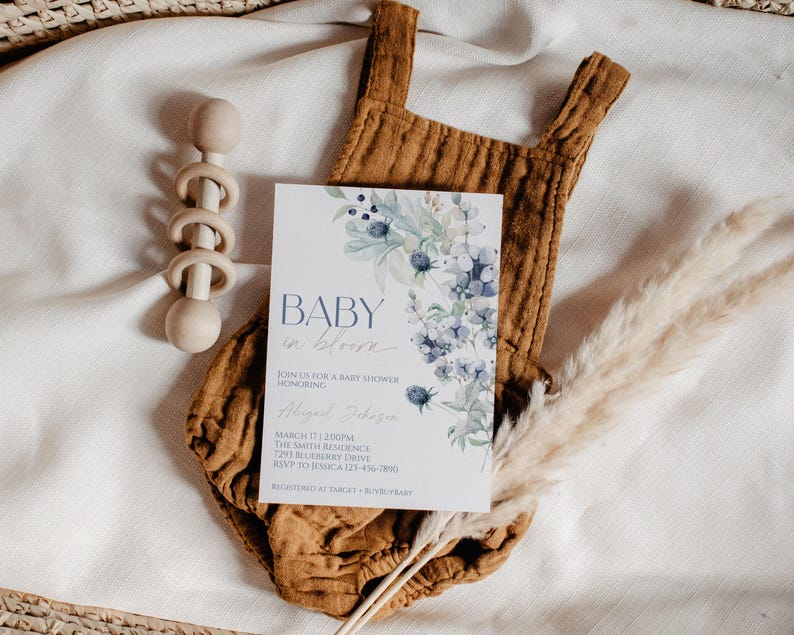 a baby shower invitation with a floral design, placed on a white cloth with baby toys and pom-poms.