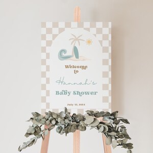 May include: A welcome sign for a baby shower with a beach theme. The sign has a checkered background and a graphic of a palm tree, surfboard, and wave. The text reads "Welcome to Hannah's Baby Shower July 13, 2024".