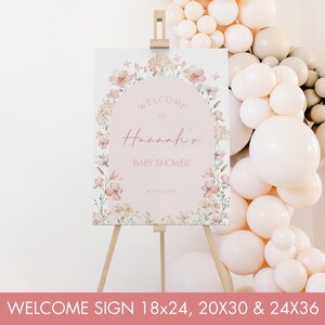 May include: A welcome sign for a baby shower featuring a floral design. The sign is pink with a white circle in the centre that says "Welcome to Hannah's Baby Shower".