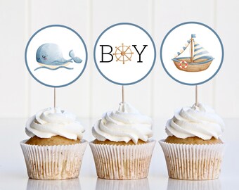 Puede incluir: Tres cupcakes decorados con adornos para cupcakes de temática náutica azul y blanca. Los adornos representan una ballena, un barco y la palabra "BOY" con un timón.