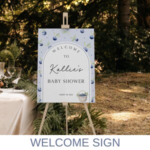 May include: A blue and white welcome sign with a watercolor blueberry design. The sign reads "Welcome to Kallie's Baby Shower, August 18, 2024".