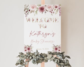 May include: A baby shower welcome sign on a wooden easel. The sign features floral accents and the text "WELCOME TO Kathryn's Baby Shower" with the date January 16, 2023. Eucalyptus greenery adorns the base of the easel.