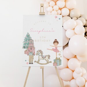 Pode incluir: Um cavalete de madeira exibe uma placa de boas-vindas para uma festa de 5º aniversário. A placa apresenta ilustrações de um quebra-nozes, um cavalo de balanço e uma bailarina. A placa é decorada com estrelas rosa e verdes e um arco de balões.
