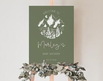 Peut inclure: Une pancarte d'accueil verte avec une illustration blanche de montagne et de forêt. Le texte indique "Bienvenue à la fête prénatale de Marley le 13 juillet 2024".