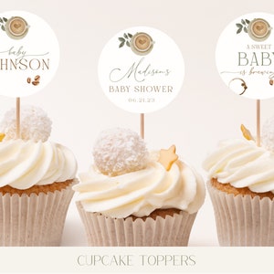 May include: Three white cupcakes with white frosting and a coffee cup themed cupcake topper. The toppers feature a coffee cup with a heart, green leaves, and brown coffee beans. The text on the toppers reads "baby Johnson", "Madison's Baby Shower 06.21.23", and "A Sweet Baby is brewing".