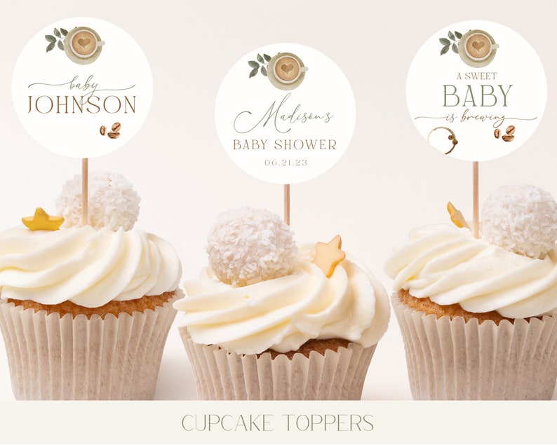May include: Three white cupcakes with white frosting and a coffee cup themed cupcake topper. The toppers feature a coffee cup with a heart, green leaves, and brown coffee beans. The text on the toppers reads "baby Johnson", "Madison's Baby Shower 06.21.23", and "A Sweet Baby is brewing".